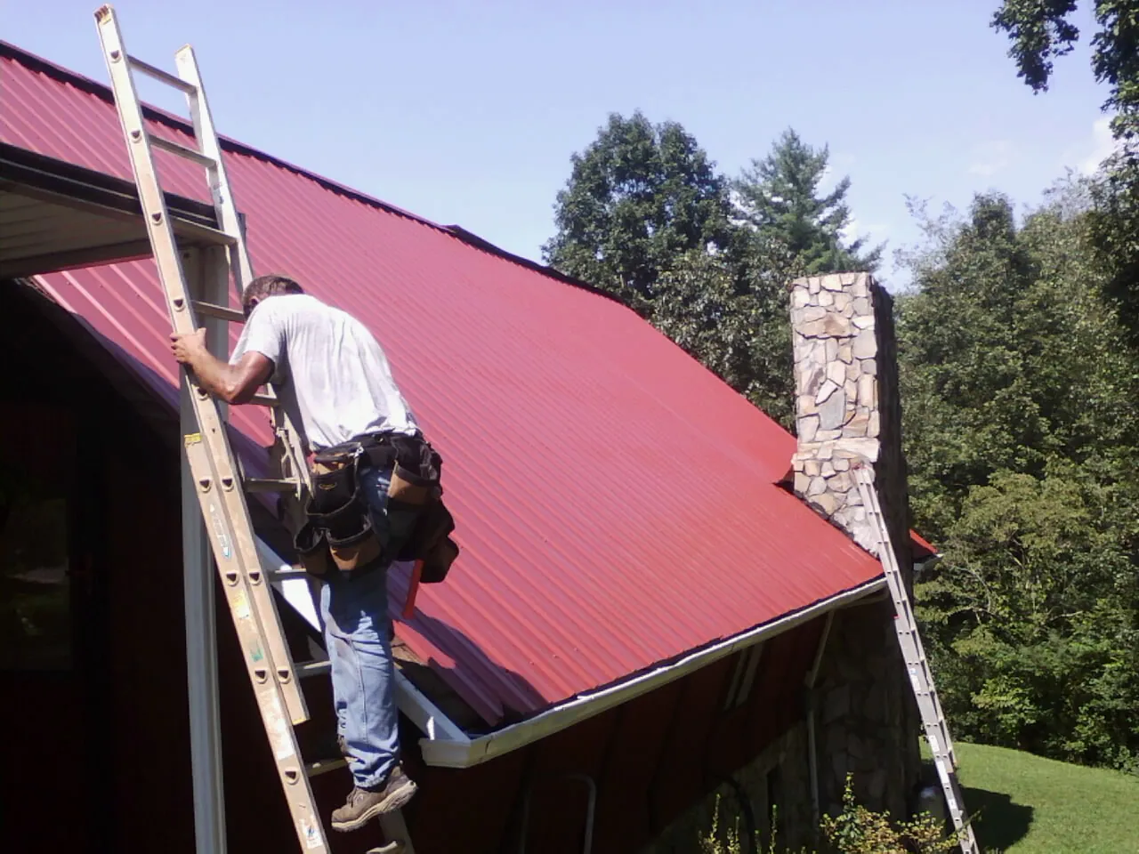 Roofer on a ladder beside a red metal roof and stone chimney — Soffit & Fascia Repair by Roofing Brothers in Spicewood